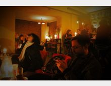 Eine gemütliche Café-Bar im warmen Kerzenlicht, mit Ziegelwänden und Holzdekor; Menschen sitzen an kleinen Tischen und hohen Barhockern, einige im Gespräch vertieft, während andere ihr Getränk genießen.