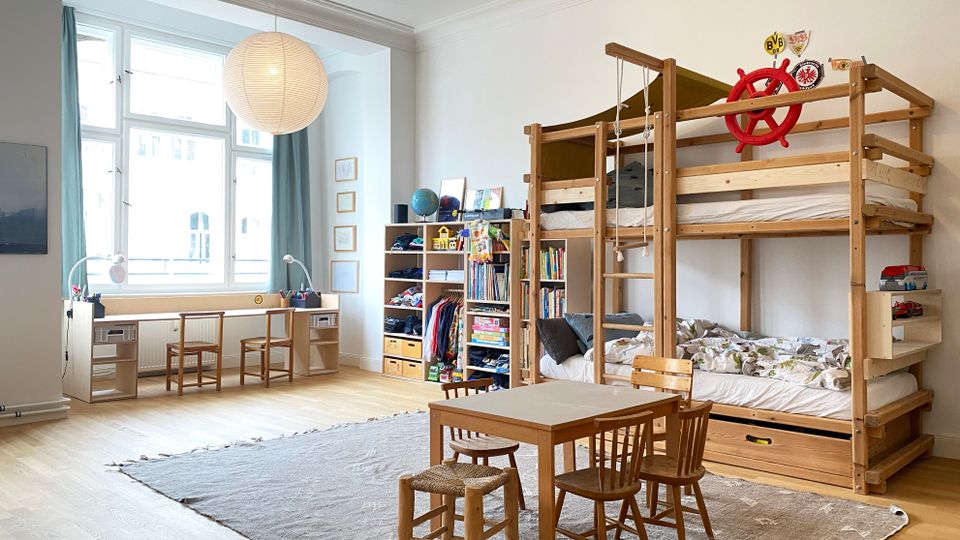 Helles Kinderzimmer mit hohen Decken und großem Fenster, einem langen Schreibtisch mit zwei Stühlen sowie Regal- und Stauraummöbeln an der Wand. Rechts steht ein großes Holz-Hochbett mit Leiter und Seilen; davor ein kleiner Tisch mit vier Stühlen auf einem Teppich.