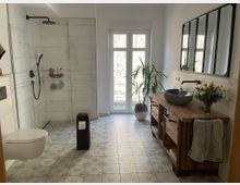 Modernes Badezimmer mit bodengleicher Dusche, weißen Fliesen und einer Holz-Waschtischkonsole mit Natursteinwaschbecken. Ein großes Fenster lässt Tageslicht ein, und Zimmerpflanzen sowie dekorative Elemente ergänzen den Raum.