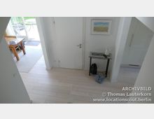 Heller Innenraum mit weiß gestrichenen Wänden, hellem Holzboden und weißen Türen. Rechts steht ein kleiner Tisch mit Deko und darunter ein Rucksack, links führt der Blick zu einem Essbereich mit einem Holztisch und Stühlen nahe einer Glastür, die zu einem Garten führt.