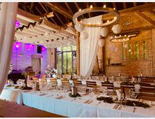 Rustikale Veranstaltungshalle mit Holzbalkendecke und sichtbaren Ziegelwänden, dekoriert für ein festliches Essen. Lange, weiß gedeckte Tische sind mit Gläsern, Besteck und schlichten, natürlichen Dekorationen gedeckt; runde Kronleuchter und hängende Papierdekorationen schmücken die Decke.