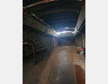 Ein langer, schmaler Lagerraum mit einer Wellblechdecke und Betonwänden. Im Raum befinden sich Holzarbeiten, verstreute Gegenstände und Werkzeuge, während eine offene Tür am Ende schwaches Licht hereinlässt.