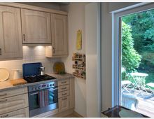 Kleine, helle Küche mit Holzfronten, integriertem Herd und Gasherdplatte, Fliesenspiegel in Weiß und einer Arbeitsplatte aus Stein. Rechts führt ein großes Fensterblick auf eine Terrasse mit weißen Gartenmöbeln und üppigem Grün im Hintergrund.