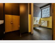 Barrierefreies Badezimmer mit gelben Fliesen an der Wand und dunklem Boden. Zu sehen sind eine Toilette mit Haltegriffen, ein Fenster mit Gitter und daneben eine getrennte Toilettenkabine.