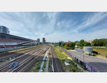 Blick auf mehrere Bahngleise, die sich durch eine urbane Umgebung erstrecken, mit der "East Side Mall" links im Bild und vereinzelten Hochhäusern im Hintergrund. Rechts sind Grünflächen, ein Parkplatz und kleinere Gebäude zu sehen, der Himmel ist leicht bewölkt.