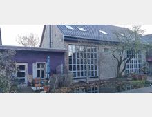 Ehemaliges Backsteingebäude mit großen Glasfenstern, einem angebauten lilafarbenen Nebenraum mit alten Holzfenstern und einem kleinen Gartenbereich mit einem Baum und einem Teich im Vordergrund.