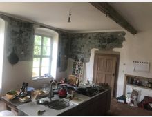 Küche mit rustikalem Charakter, geprägt durch freiliegende, teilweise verputzte Wände und eine Holzdecke mit sichtbaren Balken. Im Vordergrund steht eine Kücheninsel mit Spüle, Geschirr und Küchengeräten; durch ein großes Fenster fällt Tageslicht ein.