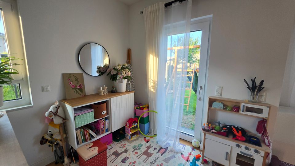 Heller Spielbereich in einem Wohnraum mit weißer Wand, niedrigem Regal mit rundem Spiegel darüber und Spielteppich auf dem Boden. Rechts führt eine verglaste Terrassentür mit transparenten Vorhängen in den Garten; daneben steht eine Kinder-Spielküche und verschiedene Spielsachen.