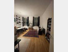 Helles Wohnzimmer mit Holzboden, zwei großen Fenstern mit dunklen Vorhängen, einem Esstisch mit Stühlen links und einem roten Teppich in der Mitte. Rechts befinden sich ein Bücherregal und eine Kommode, an der Wand hängen gerahmte Bilder.