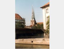 Ein Stadtbild zeigt einen Fluss im Vordergrund, eine Brücke mit Geländer, und dahinter eine Kirche mit hohem, spitzem Kirchturm. Umgeben sind die Gebäude von weiteren historischen Bauwerken und Bäumen.