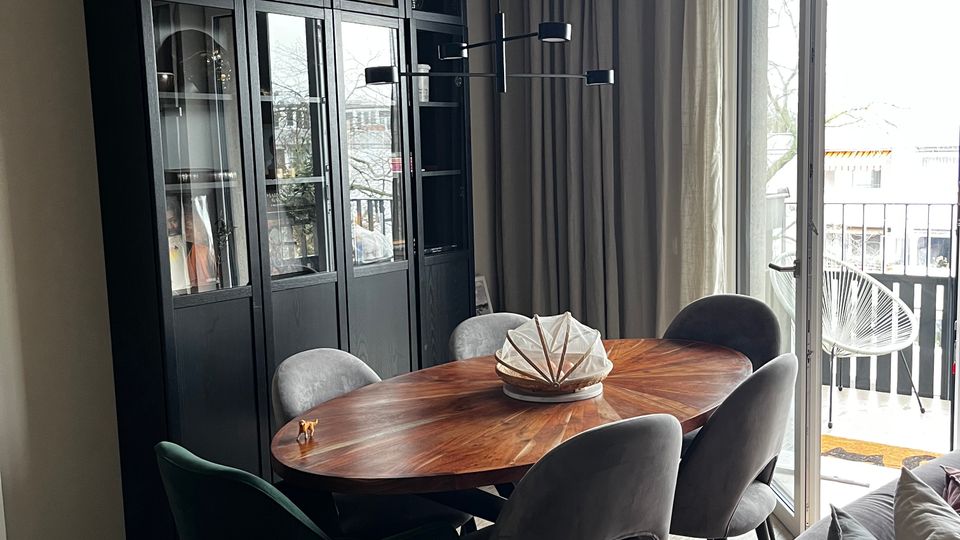 Moderner Essbereich mit ovalem Holztisch und gepolsterten Stühlen vor einem hohen, schwarzen Vitrinenschrank mit Glastüren und einer minimalistischen Pendelleuchte. Rechts öffnen bodentiefe Glastüren zum Balkon mit Vorhängen und Blick nach draußen.