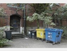 Fassade eines alten Backsteingebäudes mit einer Holztür, die über eine kleine Treppe erreichbar ist, daneben mehrere Müllcontainer in verschiedenen Farben (blau, gelb, schwarz). Im Vordergrund stehen Bäume und Büsche, die den Eingangsbereich teilweise verdecken.