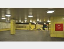 U-Bahn-Station mit gelb gefliesten Wänden, grauem Boden und beleuchtet durch runde Deckenlampen. Im Hintergrund sind Gleise, Fahrtrichtungsanzeigen und ein Bahnsteig zu sehen.