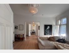 Wohnzimmer in heller Altbauwohnung mit Holzdielenboden, einer beigen Sofaecke, großen Fenstern mit halb geöffnetem Vorhang und modernem Gemälde an der Wand. Im Hintergrund ein angrenzendes Esszimmer mit geöffneten Doppeltüren und einem Tisch mit Stühlen.