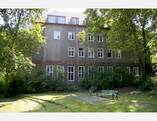 Mehrstöckiges Gebäude mit grauer Fassade und weißen Fensterrahmen, umgeben von Bäumen und Sträuchern. Im Vordergrund ein kleiner Park mit grüner Rasenfläche, einer Sitzbank und einer Mülleimer.