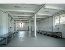 Großer, leerer industrieller Küchenraum mit weißen gefliesten Wänden und grauem Boden. Der Raum verfügt über Edelstahl-Arbeitsflächen, mehrere Wasseranschlüsse und ein großes Fenster, das Licht hereinlässt.