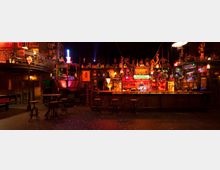 Eine stimmungsvoll beleuchtete Bar in rustikalem Design mit einer langen, hölzernen Theke, umgeben von dekorativen Objekten wie Neonlichtern, Schildern und Figuren. Im Hintergrund befinden sich Regale mit Getränken, und auf der linken Seite ist eine erhöhte Galerie mit Metallgeländer zu sehen.