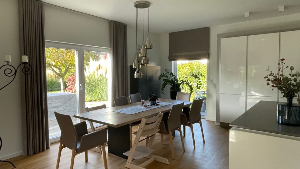 Esszimmer in einem modernen Wohnhaus mit einem großen Holztisch und sechs Stühlen. Große Fenster mit Vorhängen und Blick auf einen Garten sorgen für viel Tageslicht; links steht ein Metall-Kerzenständer, rechts eine schlichte weiße Küchenfront.