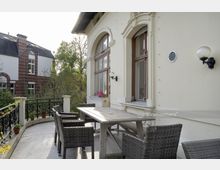 Terrasse eines historischen Gebäudes mit cremefarbener Fassade und dekorativen Fensterelementen, ausgestattet mit einem Holztisch und geflochtenen Stühlen; im Hintergrund ein weiteres Gebäude aus rotem Backstein und umliegende Bäume.