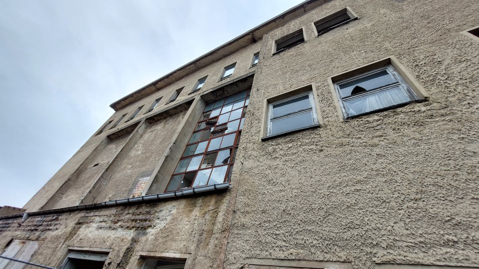Außenansicht eines alten, verputzten Gebäudes mit großen, vertikal ausgerichteten Fensterflächen und kleineren, rechteckigen Fenstern. Die Fassade wirkt verwittert und zeigt deutliche Spuren von Abnutzung.