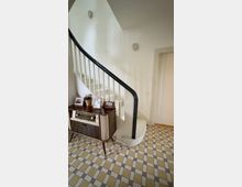 Innenflur eines Wohnhauses mit weiß gestrichener, geschwungener Treppe und dunklem Handlauf. Auf dem gefliesten Boden steht ein niedriges Sideboard mit altem Radio, Bilderrahmen und Pflanze neben einer geschlossenen Tür.