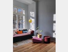 Wohnzimmer mit Holzboden und großen Fenstern, die viel Tageslicht hereinlassen. Ein Sofa mit bunten Kissen steht an der Wand, daneben ein rosa Tisch mit einer Topfpflanze und einer Lampe sowie ein kleiner roter Hocker.