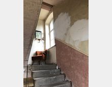 Treppenhaus in einem älteren Gebäude mit stark abgenutzten Wänden, teilweise gestrichen. Auf dem Treppenabsatz steht ein roter Polstersessel, daneben ein Fenster, das Tageslicht hereinlässt, und ein kleines, gerahmtes Bild an der Wand.