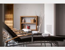 Ruheraum in einem Wellnessbereich mit mehreren Liegen, die mit zusammengerollten Handtüchern belegt sind. Im Hintergrund ein Regal mit Holz-Elementen, darauf Bücher, ein Holzeimer und eine Flasche; daneben eine schlichte Vase mit dekorativen Ästen. Links ein Eingang zu einer Sauna mit Holzverkleidung.