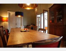 Esszimmer mit einem langen Holztisch, umgeben von Stühlen, und einer Hängelampe mit goldener Innenfläche. Im Hintergrund ein Holzbuffet, eine geöffnete Terrassentür und eine Stehlampe mit rotem Lampenschirm.