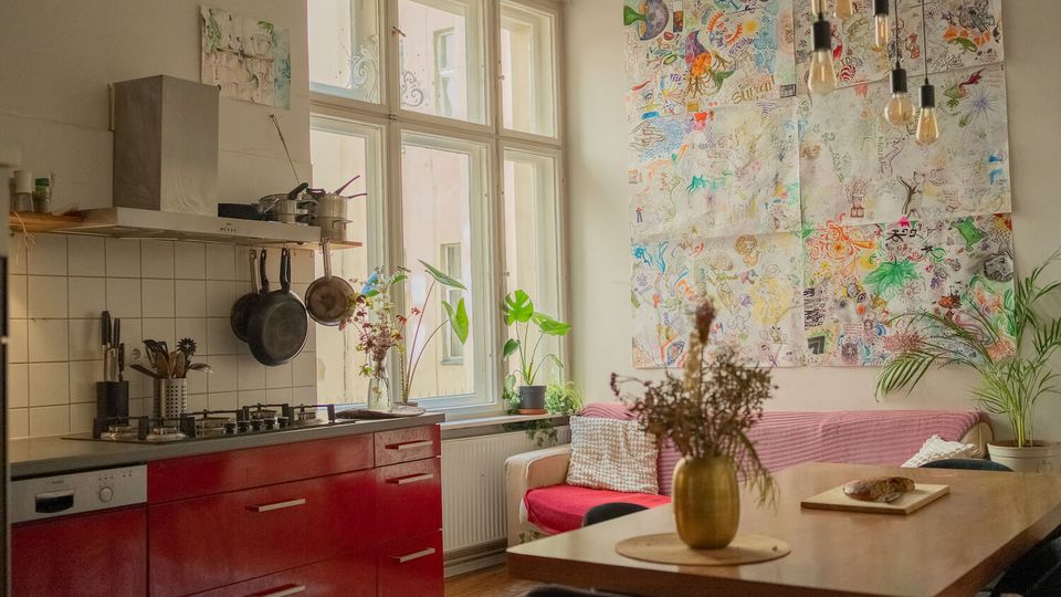 Bright open-plan kitchen and dining area with red lower cabinets, a tiled backsplash, hanging pots, and large multi-pane windows. A dining table sits beside a pink-covered sofa, with potted plants and a large wall collage of colorful drawings under hanging bulb lights.