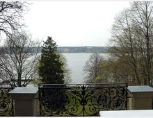 Blick von einer Terrasse mit kunstvoll verziertem Eisengeländer auf einen See, umgeben von kahlen und vereinzelt nadelgrünen Bäumen. Im Hintergrund sind das gegenüberliegende Ufer und einige Gebäude in der Ferne zu sehen.