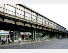 Hochbahnhof mit Metallkonstruktion, darunter ein Gehweg mit Passanten, geparkten Fahrrädern und Geschäften. Eine breite Straße verläuft im Vordergrund.
