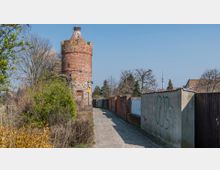 Ein gepflasterter Weg führt zu einem runden, roten Backsteinturm mit einem steinernen Sockel und einem Storchennest auf der Spitze. Der Weg ist von niedrigen Mauern und Büschen gesäumt, im Hintergrund sind Bäume und vereinzelte Gebäude sichtbar.