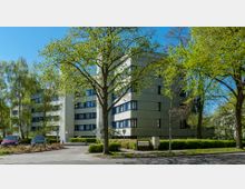 Mehrstöckiges Wohngebäude mit heller Fassade und blauen Fensterrahmen, umgeben von hohen Bäumen und gepflegten Grünflächen. Im Vordergrund sind geparkte Autos, ein Fußweg und Straßenschilder sichtbar.