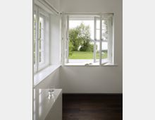 Ein heller, minimalistischer Raum mit einem großen, geöffneten Fenster, das den Blick auf eine grüne Rasenfläche mit Bäumen und einem See im Hintergrund freigibt. Im Vordergrund steht ein weißes Möbelstück mit einem Glas Wasser darauf, der Boden ist aus dunklem Holz.