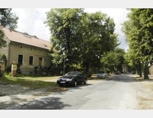 Eine ruhige Wohnstraße mit gepflastertem Gehweg, gesäumt von hohen Bäumen. Links steht ein längliches Wohnhaus mit Ziegeldach und eingezäuntem Vorgarten, während Autos am Straßenrand parken.