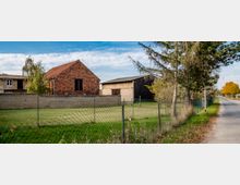 Ländlich geprägte Szenerie mit einem Backsteingebäude und mehreren Scheunen, von einem Maschendrahtzaun und einer niedrigen Mauer umgeben. Im Vordergrund Grünflächen und Bäume, eine Straße verläuft rechts am Bildrand entlang.