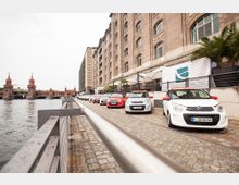 Eine gepflasterte Uferpromenade mit geparkten weißen Autos entlang eines großen Backsteingebäudes. Im Hintergrund ist die Oberbaumbrücke mit ihren markanten Türmen über der Spree sichtbar.