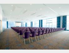 Großer, heller Konferenzraum mit Reihen von lila Stühlen, die auf eine Leinwand ausgerichtet sind. Der Raum hat einen Teppichboden, eine abgehängte Decke mit Beleuchtung und große Fensterfronten mit blauen Vorhängen.