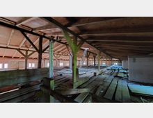 Dachboden eines Gebäudes mit Holzbalken-Konstruktion und unebenem Holzboden. Im Hintergrund sind mehrere kleine Fenster, gemauerte Wände und eine aufgestellte Leiter zu sehen.