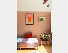 Inneneinrichtung eines Wohnzimmers mit einer rosafarbenen Wand, zwei gerahmten Bildern, einem samtigen Polsterhocker in Rosa mit einem orangefarbenen Kissen, einem Beistelltisch aus Holz mit einer Grünpflanze in einem Topf und Parkettboden. Oben rechts hängen Modelle von Planeten von der Decke.