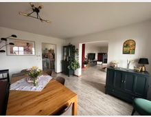 Wohnzimmer mit Essbereich in einem modernen Wohnstil. Zu sehen ist ein großer Holztisch mit Blumenschmuck, eine dunkelgrüne Kommode, helle Wände mit dekorativen Bildern, eine angrenzende offene Küche und ein Wohnbereich mit Fernseher und roten Akzentfarben.