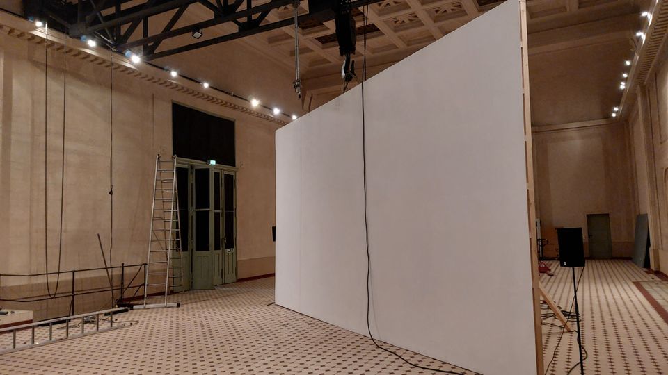 Großer Innenraum mit hohen Decken und einer modularen weißen Stellwand in der Mitte. Der Raum hat helle Wände, ein gemustertes Fliesenbodenmuster, Stahlträger an der Decke und eine grüne Doppeltür auf der linken Seite; Leitern und technische Geräte sind sichtbar.