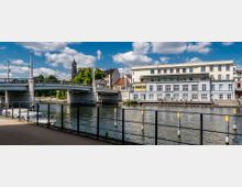 Flussufer mit einer Brücke im Vordergrund und einem mehrstöckigen Gebäude mit rechteckigen Fenstern am Wasser. Im Hintergrund ragt ein Kirchturm hinter grünen Bäumen hervor, unter einem teils bewölkten blauen Himmel.