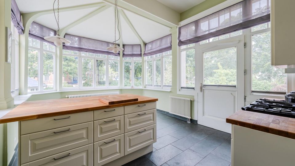 Helle Küche mit großem Kücheninselblock aus Holz und hellen Schrankfronten, dunklem Fliesenboden und Gasherd. Rundumlaufender Wintergarten-Erker mit vielen Sprossenfenstern, sichtbaren Deckenstreben, zwei Hängelampen sowie Tür nach außen.