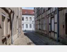 Schmale gepflasterte Gasse mit alten Gebäuden auf beiden Seiten, darunter eine Fachwerkfassade rechts und verputzte Fassaden links; im Hintergrund ein weißes Gebäude mit roten Dachziegeln und geschlossenen Fenstern sowie eine grüne Laterne in der Mitte der Gasse.