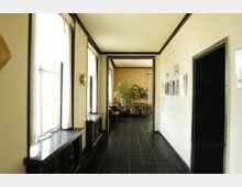 Ein heller Flur mit beigefarbenen Wänden, großen Fenstern auf der linken Seite und schwarzen Fensterbänken. Am Ende des Flurs öffnet sich ein Raum mit Sitzmöbeln und einer großen Pflanze.