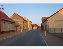 Straßenszene in einem Wohngebiet mit mehreren aneinandergebauten Häusern, die teils verputzt und teils gestrichen sind, und roten Ziegeldächern. Der Gehweg ist mit Pflastersteinen ausgelegt, und eine rote Straßenlaterne sowie kleine Vorgärten prägen die Umgebung.