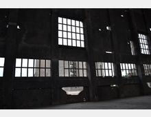 Innenraum eines verlassenen Industriegebäudes mit hohen, beschädigten Wänden aus dunklem Beton. Große Fenster mit zerbrochenen Scheiben lassen Tageslicht hinein, während Teile der Mauer unter den Fenstern eingestürzt sind.