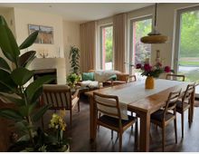 Heller Wohn- und Essbereich mit Holzesstisch und Stühlen, Ledersofa neben einem Kamin sowie mehreren Zimmerpflanzen. Große bodentiefe Fenster mit Vorhängen öffnen den Blick ins Grüne und auf eine Terrasse mit Wasserfläche.